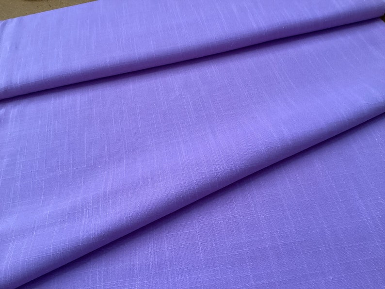 May include: A close-up of a folded piece of lavender fabric. The fabric has a subtle, textured weave, and the folds create shadows that add depth. The color is a soft, muted purple, and the fabric appears to be lightweight.