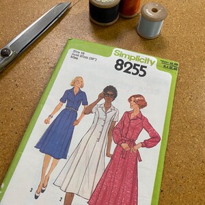 May include: A dressmaking pattern, Simplicity 8255, with illustrations of the finished dresses. The pattern is size 16, bust 97cm. Three spools of thread in black, orange, and light blue, and a pair of scissors are also visible.
