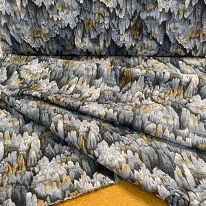 May include: A roll of fabric with a unique pattern resembling a rock formation. The design features shades of gray, white, and gold, creating a textured, almost three-dimensional effect. The fabric is folded, showcasing the pattern's repeating nature.
