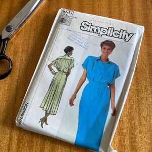 May include: A vintage Simplicity sewing pattern packet featuring a blue dress design. The packet includes illustrations of the dress and the Simplicity logo. Also visible are scissors and spools of thread.