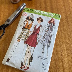 May include: A vintage Simplicity sewing pattern, number 8930, with illustrations of dresses. The packet is white with green and black text. Scissors and spools of thread are also visible, indicating a sewing theme.