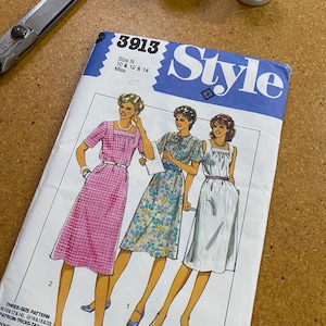May include: A vintage sewing pattern labelled "Style" with the number 3913, showing illustrations of three dresses. The pattern includes sizes 10, 12, and 14. Scissors and spools of thread are in the background.