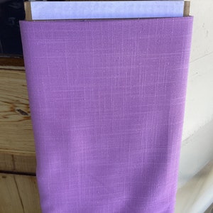 May include: A roll of solid lavender fabric with a textured weave. The fabric is tightly rolled and appears to be made of linen or a similar material. The color is a soft, muted purple, suitable for various sewing projects.