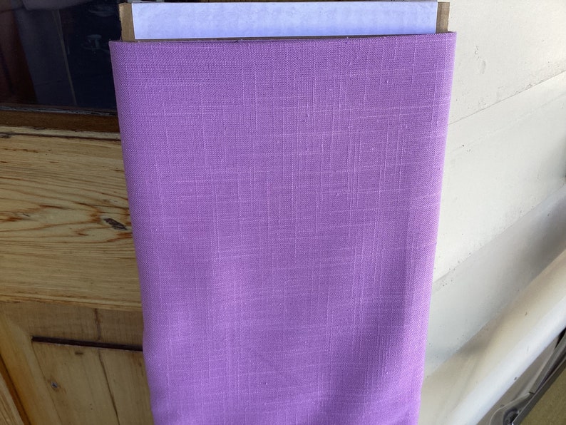 May include: A roll of solid lavender fabric with a textured weave. The fabric is tightly rolled and appears to be made of linen or a similar material. The color is a soft, muted purple, suitable for various sewing projects.