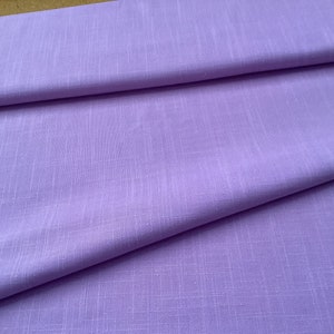 May include: A stack of folded lavender fabric. The fabric has a subtle woven texture and a soft, matte finish. The folds create shadows and depth, highlighting the fabric's drape and color.