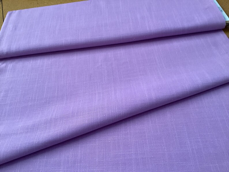 May include: A stack of folded lavender fabric. The fabric has a subtle woven texture and a soft, matte finish. The folds create shadows and depth, highlighting the fabric's drape and color.