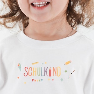 May include: A white t-shirt with a colorful graphic that says "SCHULKIND" with a rainbow design and other school-related icons.