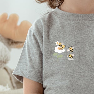 Może przedstawiać: Szara koszulka z uroczym wzorem pszczół i kwiatów. Pszczoły są żółte i czarne, a kwiat jest biały z żółtym środkiem.