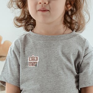 Könnte beinhalten: Ein junges Kind trägt ein graues T-Shirt mit einem kleinen Lebkuchenhaus-Motiv auf der Vorderseite. Das Kind trägt einen weißen Tüllrock und hat lange braune Haare.