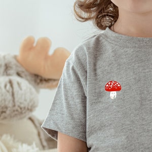 Op de afbeelding: Een grijs T-shirt met korte mouwen met een rode en witte paddenstoel afbeelding.