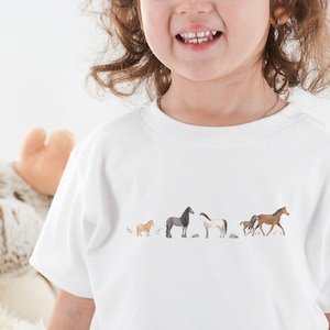 Könnte beinhalten: Ein weißes T-Shirt mit einer Reihe von fünf Pferden, die auf der Vorderseite aufgedruckt sind. Die Pferde sind braun, schwarz, weiß und beige.