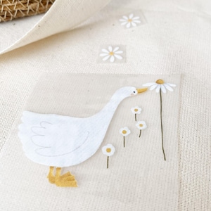 Può includere: Un'oca bianca con le zampe gialle sta annusando una margherita bianca con un centro giallo. L'oca è circondata da margherite bianche più piccole con centri gialli.