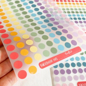 Può includere: Fogli di adesivi a pois colorati in un arcobaleno di tonalità, tra cui rosso, arancione, giallo, verde, blu e viola. Gli adesivi sono disposti in file e colonne ordinate. Il testo "BÜGELBILDER DOTS" è visibile su un'etichetta rossa.
