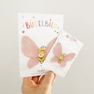 Può includere: Due toppe da stirare con una farfalla rosa e un'ape gialla che tiene una margherita. La toppa più grande dice "Bügelbild" e quella più piccola dice "Bügelbild Mini".