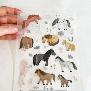 Può includere: Un foglio di adesivi di cavalli colorati con una varietà di pose e design. Gli adesivi sono su uno sfondo bianco con un bordo grigio chiaro. La parola "Pferde" è stampata nella parte inferiore del foglio.