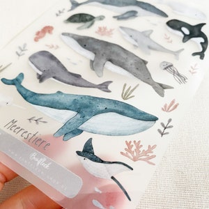 May include: A sheet of clear stickers featuring watercolor illustrations of various sea creatures, including whales, dolphins, a sea turtle, a manta ray, a jellyfish, and a seahorse. The stickers are labeled "Meerestiere" and "Buntfleck".