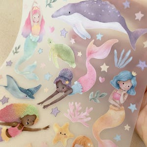 Könnte beinhalten: Ein transparenter Aufkleberbogen mit Aquarellillustrationen von drei Meerjungfrauen mit unterschiedlichen Haarfarben, einem großen lila Wal, einer grünen Meeresschildkröte, einem blauen Rochen, einem gelben Seestern, rosa Korallen sowie kleinen pastellfarbenen Sternen und Herzen.
