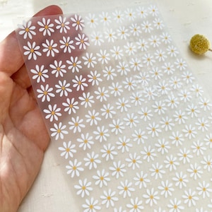 Könnte beinhalten: Ein Blatt durchsichtiges Klebepapier mit einem sich wiederholenden Muster aus kleinen weißen Gänseblümchen mit gelben Zentren.