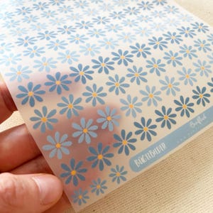 Könnte beinhalten: Ein transparenter Bogen mit Bügelbildern, der ein wiederkehrendes Muster aus kleinen blauen Gänseblümchen mit gelben Mitten zeigt. Unten rechts befindet sich ein blaues rechteckiges Etikett mit dem Aufdruck Buntfleck und BÜGELBILDER.