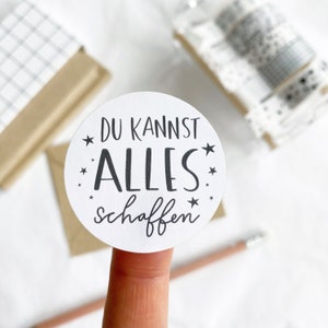 Könnte beinhalten: Ein weißer runder Aufkleber mit dem deutschen Text "DU KANNST ALLES schaffen" in Schwarz, umgeben von kleinen schwarzen Sternen. Der Aufkleber wird von einem Finger gehalten. Weitere Artikel im Hintergrund sind ein Bleistift und Schreibwaren.