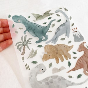 Könnte beinhalten: Eine Blatt mit bunten Dinosaurier-Stickern mit verschiedenen Dinosauriern, darunter ein Triceratops, ein Stegosaurus, ein Pterodactylus und ein T-Rex. Die Sticker sind auf weißem Hintergrund und perfekt, um jedem Projekt eine lustige und fantasievolle Note zu verleihen.
