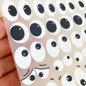 Könnte beinhalten: Blatt mit weißen und schwarzen Wackelaugen-Aufklebern in verschiedenen Größen. Die Augen haben schwarze Pupillen und weiße Iris. Einige haben einen Cartoon-Ausdruck. Die Aufkleber befinden sich auf einer transparenten Folie.