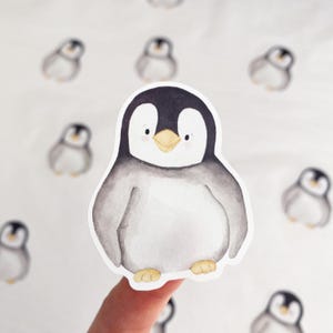 Puede incluir: Una pegatina de acuarela de un pingüino de dibujos animados. El pingüino tiene una cabeza negra, un cuerpo gris y un pico y patas amarillas. Hay varias otras pegatinas de pingüinos en el fondo. El pingüino tiene una expresión amigable y sonriente.