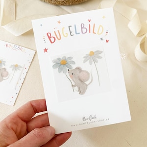 Könnte beinhalten: Eine weiße Karte mit dem Wort "BÜGELBILD" in bunten Buchstaben, die eine graue Maus mit einer Gänseblume zeigt. Die Karte trägt auch den Text "Buntfleck" und "www.buntfleck-shop.de".