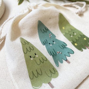 Könnte beinhalten: Drei kleine weiße Stoffgeschenktüten mit Kordelzug. Jede Tasche zeigt einen anderen Weihnachtsbaum im Cartoon-Stil mit einem lächelnden Gesicht. Ein Baum ist grün, einer ist blau und einer ist grün mit goldenen Sternen.