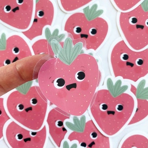 Könnte beinhalten: Ein Set süßer, rosa Erdbeer-Aufkleber mit fröhlichen Gesichtern und grünen Blättern. Die Aufkleber sind transparent und haben eine glänzende Oberfläche.