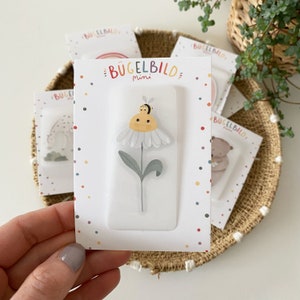 Può includere: Una carta bianca con pois colorati e il testo "Bügelbild Mini" stampato su di essa. La carta presenta una finestra di plastica trasparente che mostra una margherita bianca con un centro giallo e una piccola ape in cima.
