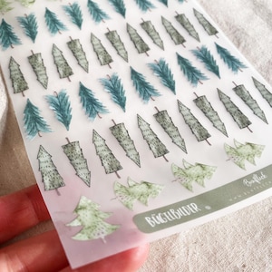 May include: A sheet of iron-on transfers featuring various watercolor-style evergreen tree designs in shades of green and blue. The sheet includes the text "BÜGELBILDER".