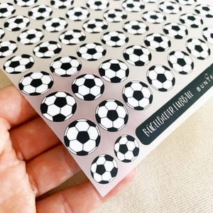 Pode incluir: Uma folha com múltiplas transferências circulares termocolantes apresentando um padrão clássico de bola de futebol em preto e branco. A folha inclui gráficos de bolas de vários tamanhos e uma etiqueta preta com o texto branco BÜGELBILDER FUSSBALL.
