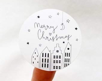 Puede incluir: Pegatina redonda blanca con texto negro que dice "Feliz Navidad" con un símbolo de corazón y una luna creciente. La pegatina también tiene una ilustración en blanco y negro de un pueblo pequeño con casas y estrellas.