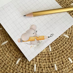 Könnte beinhalten: Ein weißes Blatt kariertes Papier mit einer Aquarellillustration einer kleinen grauen Maus, die einen gelben Stift hält. Ein goldfarbener Stift liegt diagonal auf dem oberen Teil des Papiers und zeigt auf die Mauszeichnung.