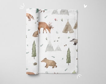 May include: A white muslin baby blanket with a woodland animal print. The print features deer, bears, foxes, badgers, trees, mountains, and teepees. The blanket is soft and absorbent, making it perfect for cuddling and comforting your little one.
