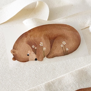 Könnte beinhalten: Illustration eines braunen Bären mit weißen Gänseblümchen-Akzenten. Der Bär ist zusammengerollt, der Kopf ruht auf seinen Pfoten. Das Kunstwerk befindet sich auf weißem Hintergrund, was auf einen Druck oder Aufkleber hindeutet.