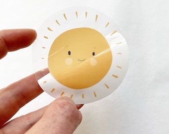 Könnte beinhalten: Ein transparenter Aufkleber mit einer gelben Sonne mit einem lächelnden Gesicht und weißen Strahlen.