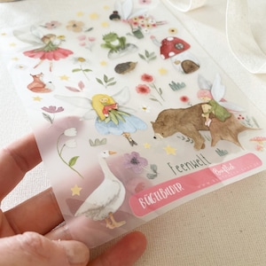 May include: A sheet of iron-on transfer stickers featuring a whimsical fairy world theme. The stickers include fairies, a goose, a bear, a frog, a hedgehog, a mushroom house, and flowers. The sheet is labeled "Feenwelt" and "Bügelbilder".