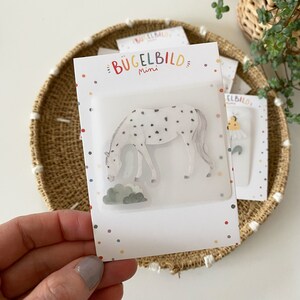 Könnte beinhalten: Eine weiße Karte mit schwarzen Punkten an den Rändern. Die Karte trägt den Text "Bügelbild mini" in bunten Buchstaben. Die Karte zeigt einen transparenten Aufkleber eines weißen Pferdes mit schwarzen Flecken, das auf grünem Gras grast.