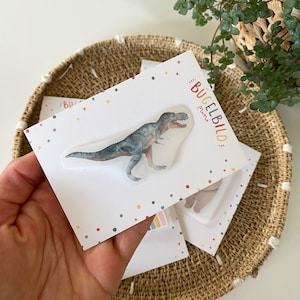 Könnte beinhalten: Eine weiße Karte mit bunten Konfettipunkten und dem aufgedruckten Text "Bügelbild mini". Auf der Karte befindet sich ein transparenter Bügelbildaufkleber mit einem grünen und grauen Dinosaurier.