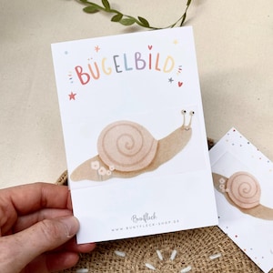 Può includere: Biglietto bianco con un'illustrazione ad acquerello di una lumaca e la scritta "BÜGELBILD" in lettere colorate. La lumaca è marrone chiaro con un guscio a spirale e piccoli fiori. Il biglietto riporta anche l'indirizzo del sito web "WWW.BUNTFLECK-SHOP.DE".