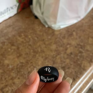 May include: A black silicone ring engraved with the letter "R" and the words "My honey" in white.
