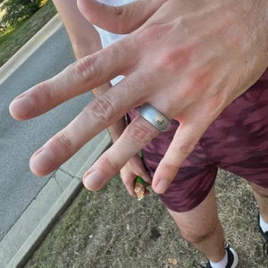 May include: A silver silicone ring with a small crescent moon design on the top. The ring is worn on the ring finger of a person's left hand.