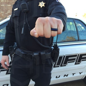 May include: A law enforcement officer wearing a black uniform with a sheriff's star badge and a black silicone ring with a thin blue line on the ring. The officer is standing next to a white sheriff's patrol car.