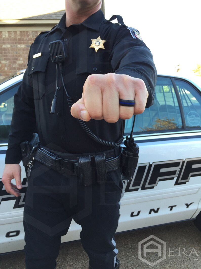 May include: A law enforcement officer wearing a black uniform with a sheriff's star badge and a black silicone ring with a thin blue line on the ring. The officer is standing next to a white sheriff's patrol car.