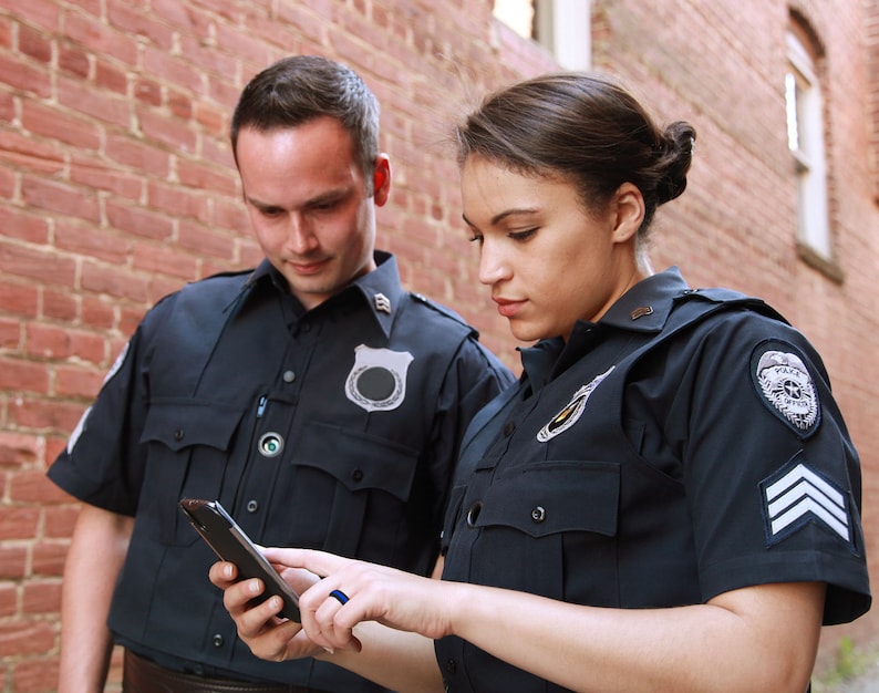May include: Two police officers in uniform, one looking at a smartphone, the other looking at the phone screen.