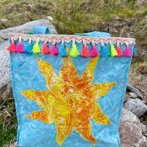 May include: A light blue tote bag featuring a large, central sun applique with marbled yellow and orange fabric. The top edge is decorated with a row of small, colorful fabric tassels in shades of pink, blue, and yellow, attached to a trim.