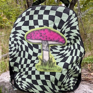 Puede incluir: Una mochila con un patrón de tablero de ajedrez distorsionado en negro y verde claro. Centrado en la parte frontal hay un gran aplique difuso de un hongo con un sombrero rosa brillante, un tallo blanco y una base verde, bordeado con costuras verde claro.