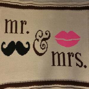 May include: A crocheted blanket in cream with brown trim and lettering. The blanket features the words "mr. & mrs." along with a black mustache and pink lips. The blanket is a handmade item.